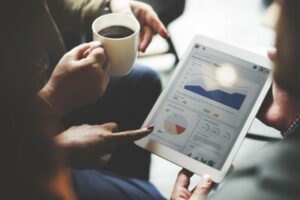 Two people review business charts on a tablet while one holds a coffee cup, engaged in discussion.