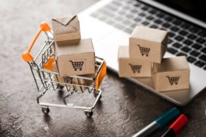 A shopping cart filled with cardboard boxes sits next to a laptop, symbolizing online shopping and e-commerce.