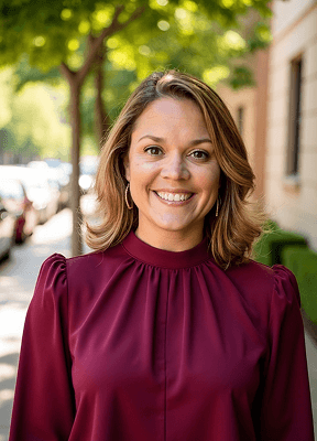 A woman with wavy hair stands outdoors in a burgundy blouse, surrounded by lush green trees and a sunny street scene.